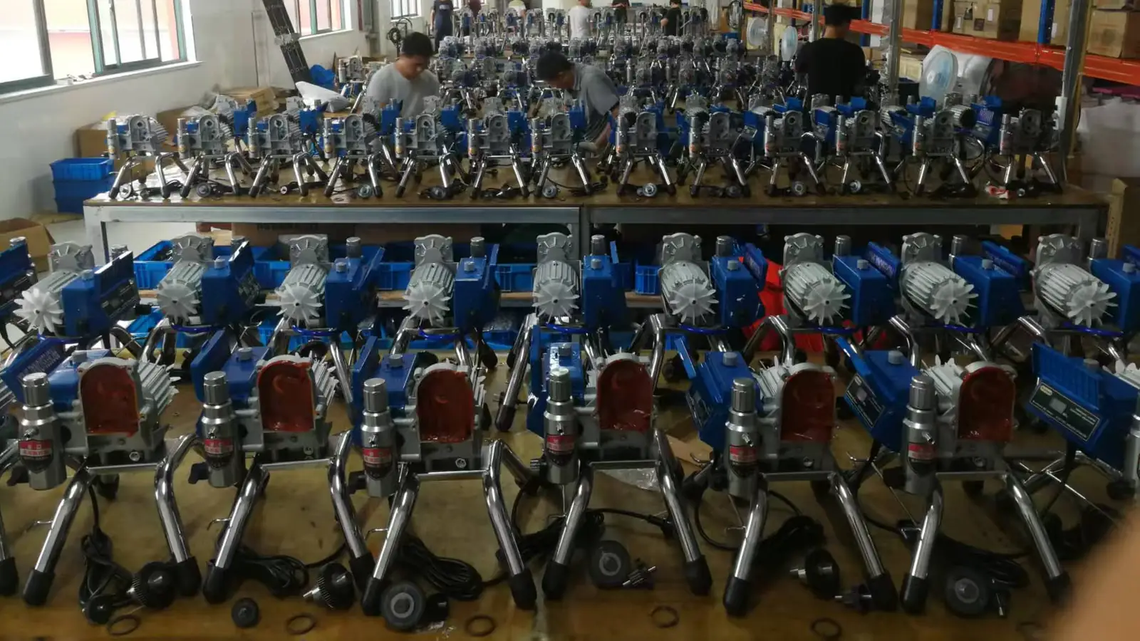 Rows of GUTUBAO airless sprayers on factory assembly benches, drive motors and pump sections in view, with workers assembling more units behind them.